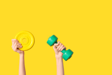 Female hands with barbell plate and dumbbell on yellow background, closeup