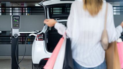 Young woman travel with EV electric car to shopping center parking lot charging in downtown city...