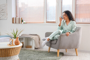 Young African-American woman using asthma inhaler in in armchair at home