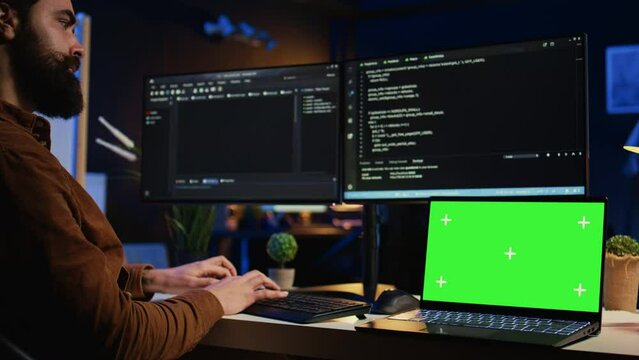 Programmer coding on computer next to isolated screen laptop in home office, developing software application. Teleworking IT specialist doing programming on PC near chroma key notebook, camera B