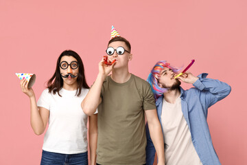 Young friends in funny disguise with party whistles on pink background. April fool's day celebration - obrazy, fototapety, plakaty