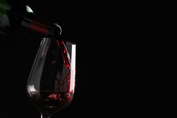 Pouring tasty red wine from bottle into glass on black background, closeup. Space for text