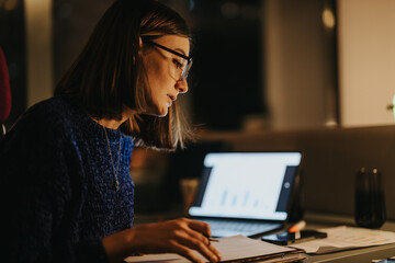 Female business executive analyzing graphs on a laptop in a modern office setting, showcasing dedication and concentration.
