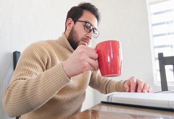 Joven apuesto con barba y anteojos sentado, tomando café mientras lee un libro en casa. Tiempo de...