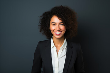 Naklejka na ściany i meble A businesswoman with curly hair, smiling warmly, wearing a black blazer over a white shirt against a dark background, conveying a professional and approachable demeanor.