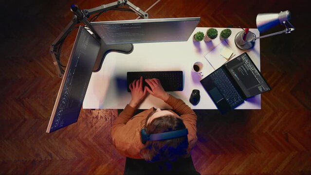 Top down view of developer typing complex code in home office, listening music and developing software application. IT expert wearing headphones, solving coding project tasks on laptop, aerial shot