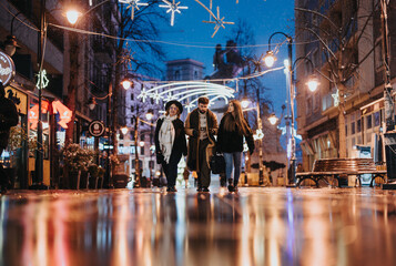 A group of young adults strolls down a wet city street adorned with festive lights, reflecting the...