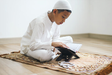Young Asian muslim boy wear skullcap, sitting on prayer mat and reading Quran
