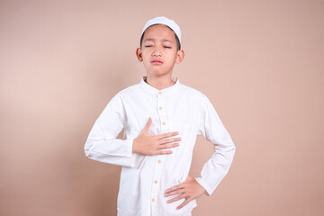Young Asian moslem boy in skullcap touching his stomach feeling hungry on fasting day 