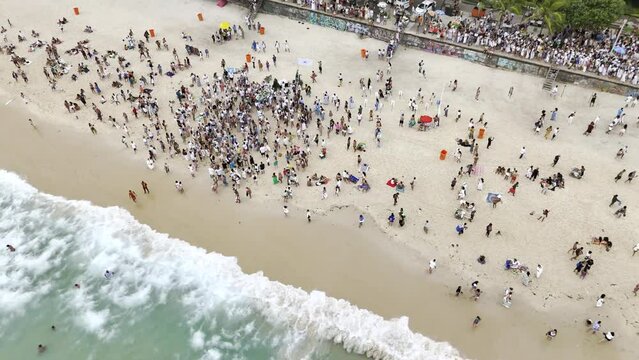 Aerial shot drone orbits high to the left over crowd as they walk into the sea by Arpoador in Ipanema, Brazil