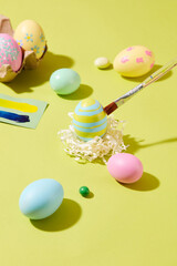 A paintbrush is painting an Easter egg placed on kraft paper with blue and yellow color. Other eggs with different colors and patterns are decorated on the surface. For Easter Day concept