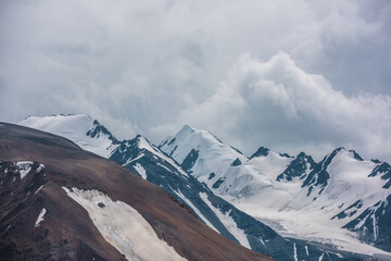 Dramatic view to large rock mountain range with snow-capped pinnacle in gray cloudy sky. Atmospheric landscape with snow cornice on snow-white pointy peak. Big rocky mountains and snowy peaked top. © Daniil