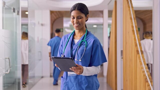 Camera tracks female doctor or nurse wearing scrubs using digital tablet in busy hospital corridor with colleagues in background - shot in slow motion