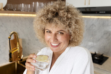 Joyful woman in white bathrobe holding a glass of water with lemon, smiling in a modern kitchen, promoting healthy lifestyle and hydration. - 743477308