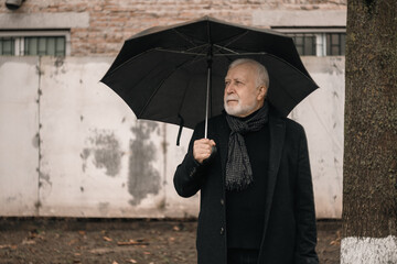 An elderly man in a black coat with an umbrella stands outside in the rain near a white fence, rainy weather