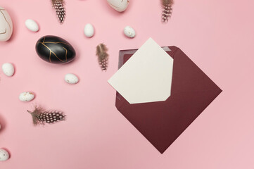 Top view of easter eggs, feathers, red envelope and white card on pink background. Creative easter composition, spring, flat lay, copy space.