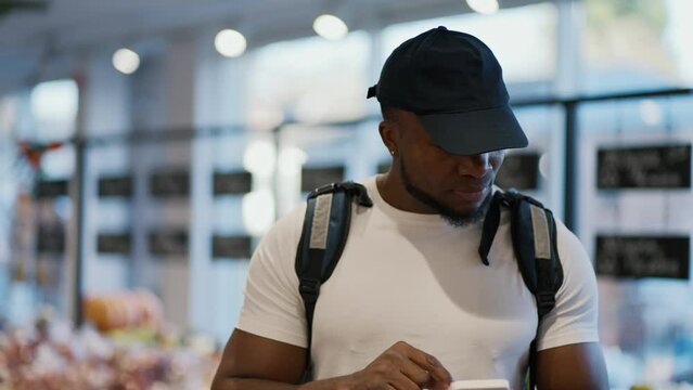 A Black man in a white T-shirt carries a large bag on his shoulders and looks at his smartphone to check the necessary products. A delivery man walks along the food counters in a grocery store and
