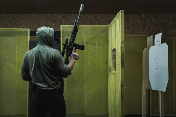 A man in a hood is engaged in tactical shooting with rifle at a range, photo from the back.