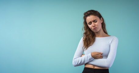 Woman holding her stomach in pain, her face grimacing with discomfort, against a solid light blue background, expressing the distress caused by stomachache - obrazy, fototapety, plakaty
