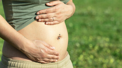 Pregnant woman with navel piercing rubbing her belly with her hands in the first weeks of pregnancy outdoors with blurred background of flowers in spring