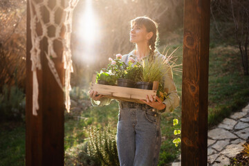 Young happy woman with a box of plants and flowers for seedlings in the garden at sunset. Flower seedlings for the garden, floriculture - 743609955