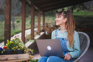 Young happy woman in denim overalls sitting with a laptop on the terrace of a country house on a spring day - 743611947