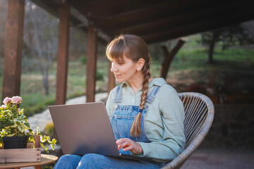 Young happy woman in denim overalls sitting with a laptop on the terrace of a country house on a spring day - 743612347