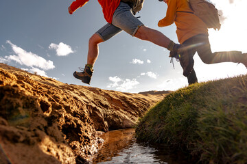 Two active young tourists hikers with backpacks jumps over a creek. Grass and sandy creek banks