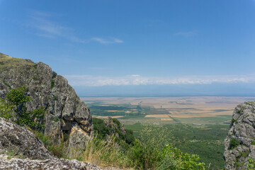 View of the Alazani Valley from a high mountain. Rocks and trees around. Agrarian fields are visible. Clear blue sky and clouds over the Caucasus ridge.