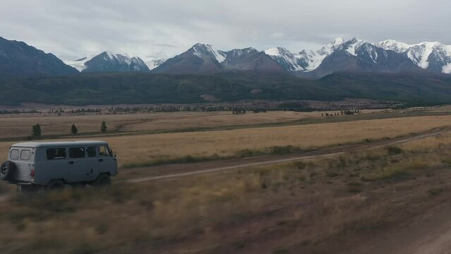 Microbus car dynamic riding meadow dirt road with dust surrounded cinematic alpine valley drone close follow shot. Automobile transportation movement sand path beautiful cliff rock scenery travel trip