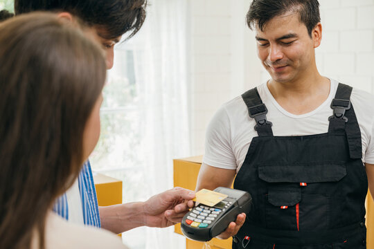 Family pays via credit card post moving. Delivery worker in uniform holds a box. Professional movers ensure efficient home relocation. Teamwork and relocation service evident. Moving Day