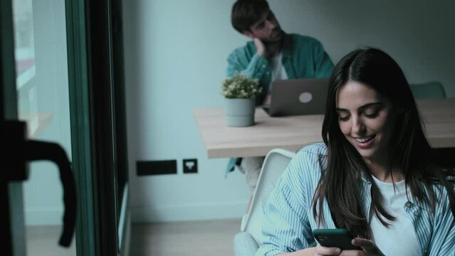 One couple young adult man and woman enjoying time at home sitting in separate places using phone and laptop. Modern millennial lifestyle indoor leisure activity. One female chatting on phone smiling