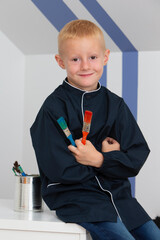 A little blond boy sitting on a desk with paintbrushes in his hands