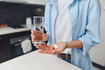 Woman taking medication and staying hydrated in the kitchen with a glass of water and pill in hand