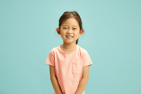 Cheerful portrait of elementary ages Asian girl radiates with a bright smile, expression joyful and happy dressed in a soft peach t-shirt standing against blue isolated background.