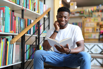 In the university library, a young man sits among bookshelves, smiling as he studies, embracing...