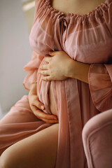Pregnant woman in a stylish pink peignoir sitting on the chair and holding her belly. Happy...
