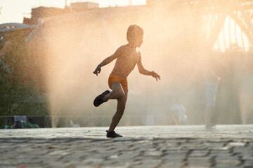 Captured in the warm glow of sunset, a joyful child dances in the water mist of an urban fountain, embodying the spirit of summer