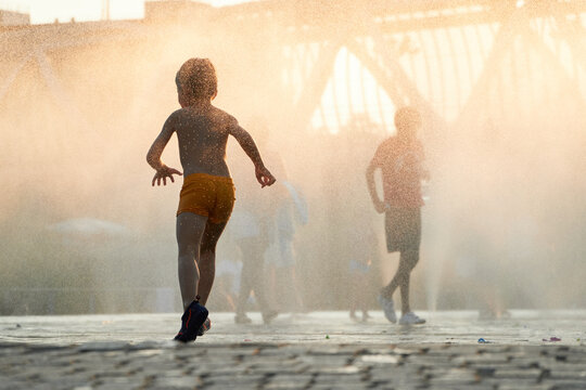Anonymous silhouette of a child runs through the mist of a city fountain as the sun descends, casting a warm, golden glow over the urban landscape