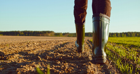 Low angle: man walking in rubber boots in a farmer's field, the blue sky above the horizon. Man walking through an agricultural field. Farmer walks through a plowed field in early spring. - 743754990
