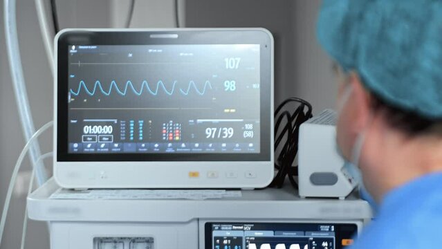 View of the monitor in the operating room, displaying the vital signs of the patient. A young surgeon is performing a laparoscopic surgery.