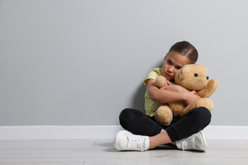 Child abuse. Upset girl with toy sitting on floor near grey wall, space for text