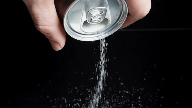 A male hand holds a soda can on a black background and pours sugar from it. Concept of harmful drinks.