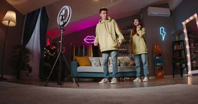 Guy and girl are filming new video for social networks, synchronized dance movements are filmed by smartphone mounted on ring lamp on tripod. Young people record dance videos for their channel. AD.