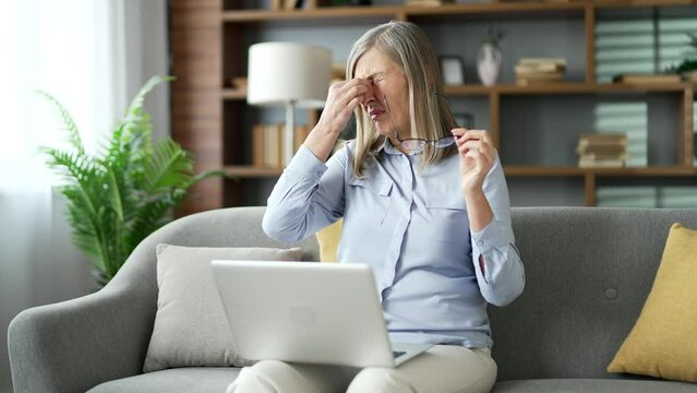 Senior female suffering from eye strain and headache working on laptop computer sitting on the sofa in the living room at home. Tired elderly woman takes off glasses and massages, rubs bridge of nose