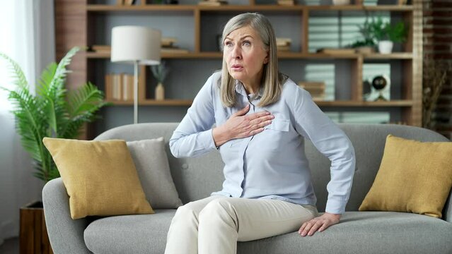Sick senior gray haired female suffering from heart attack chest tension sitting on sofa in living room at home. Elderly retired woman holding her chest with her hands, needs urgent medical attention