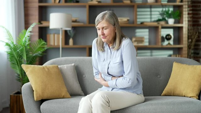 Senior female with gray hair feels stomach pain sitting on sofa in living room at home. Sick elderly retired woman has heartburn, gastritis or poisoning. Suffers from spasms, constipation or bloating