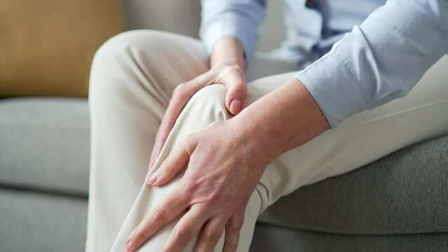 Senior female hands massaging knee leg muscles sitting on sofa in living room at home. Elderly woman suffers from painful severe knee joint pain, could not walk, illness cramps, rheumatism. Close up