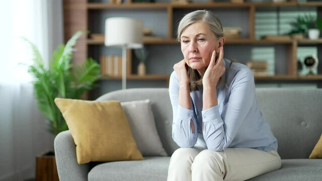 Senior gray haired female has depression suffering from mental pain sitting on the sofa in living room at home. Sad elderly retired woman has personal problems, worries, feels loneliness and boredom