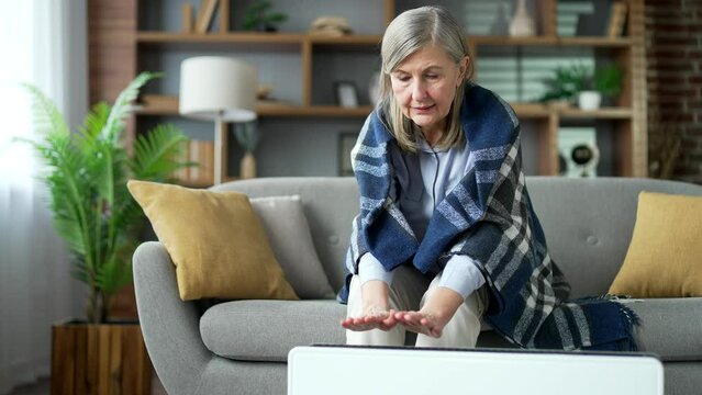 Senior gray haired female covered with blanket frozen sitting on sofa in living room at home. Upset elderly retired woman cold indoors. Grandmother feels discomfort due to low temperature in apartment